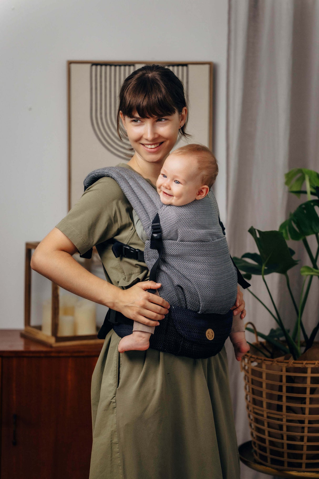 A parent wearing the LennyLamb LennyLight - Little Herringbone Ombre Grey baby carrier, smiling while holding a happy baby.| Babymaxi