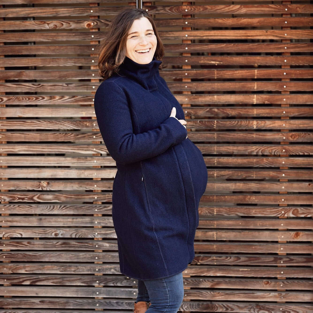 Pregnant woman wearing the Mamalila Eco Wool Babywearing Coat Oslo - Navy, showcasing its stylish design against a wooden backdrop | Babymaxi