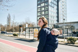 Mamalila Dublin Babywearing Coat – Navy featuring a woman with a baby on her back in a city | Babymaxi