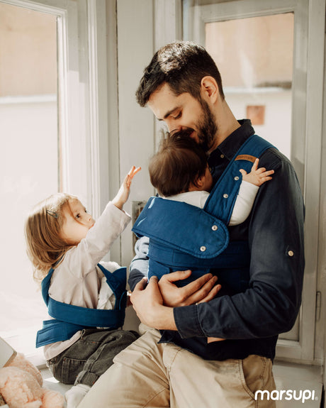 Father wearing Marsupi Classic - Ocean Blue baby carrier with two kids in a bright room | Babymaxi